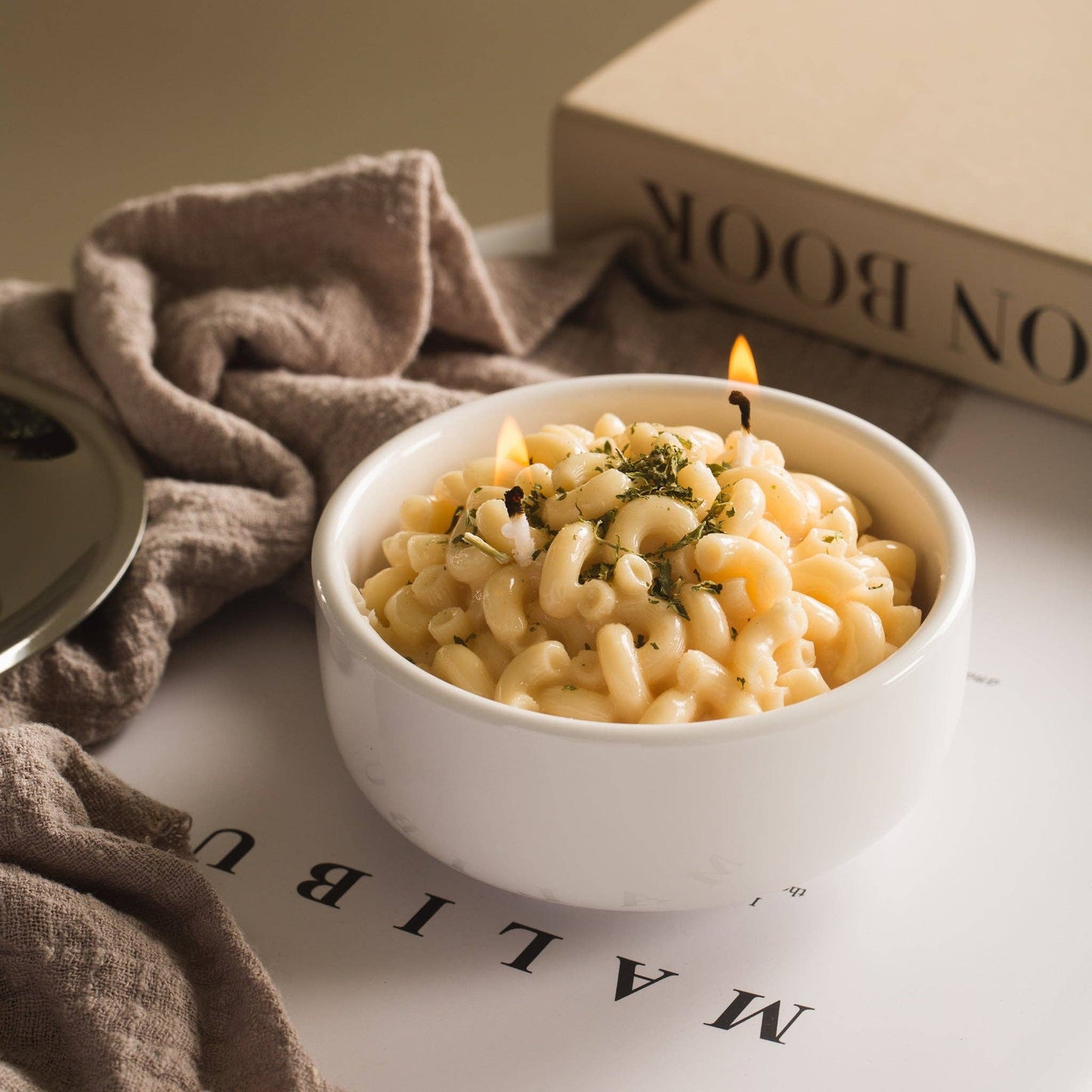 Candle that looks like a realistic bowl of white cheddar mac and cheese in a white porcelain bowl. Backdrop is a computer desk with magazines and a dish towel. Candle is lit.