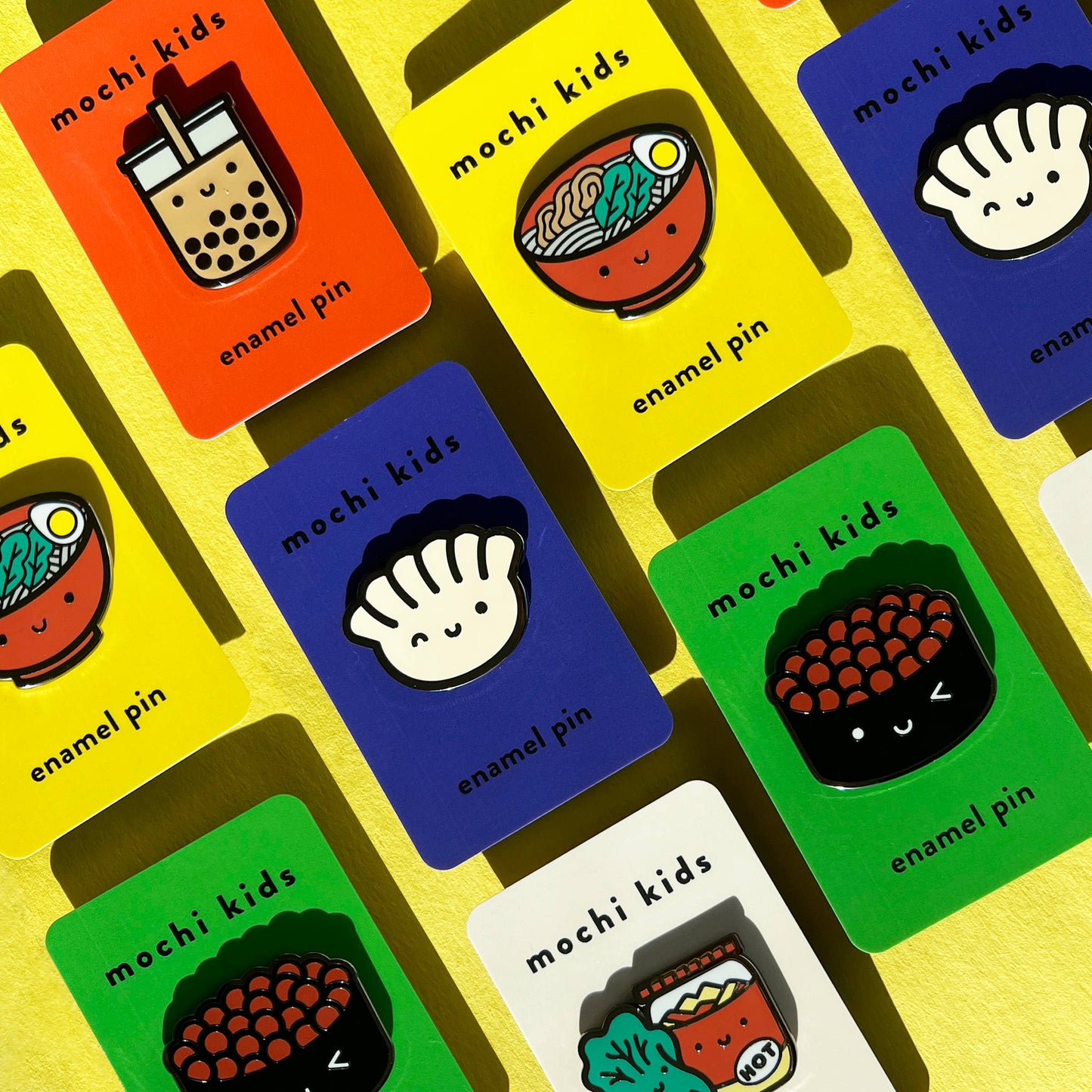 Collection of colorful packaging for 'mochi kids' enamel pins on a yellow background.