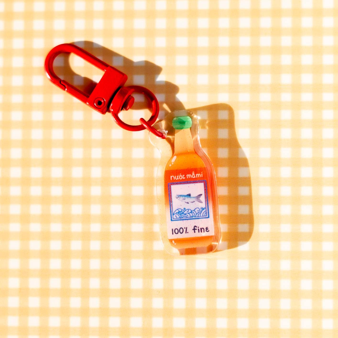 Decorative charm shaped like a bottle of fish sauce with green lid and text "nuoc mami... 100% fine" on label with picture of a fish.