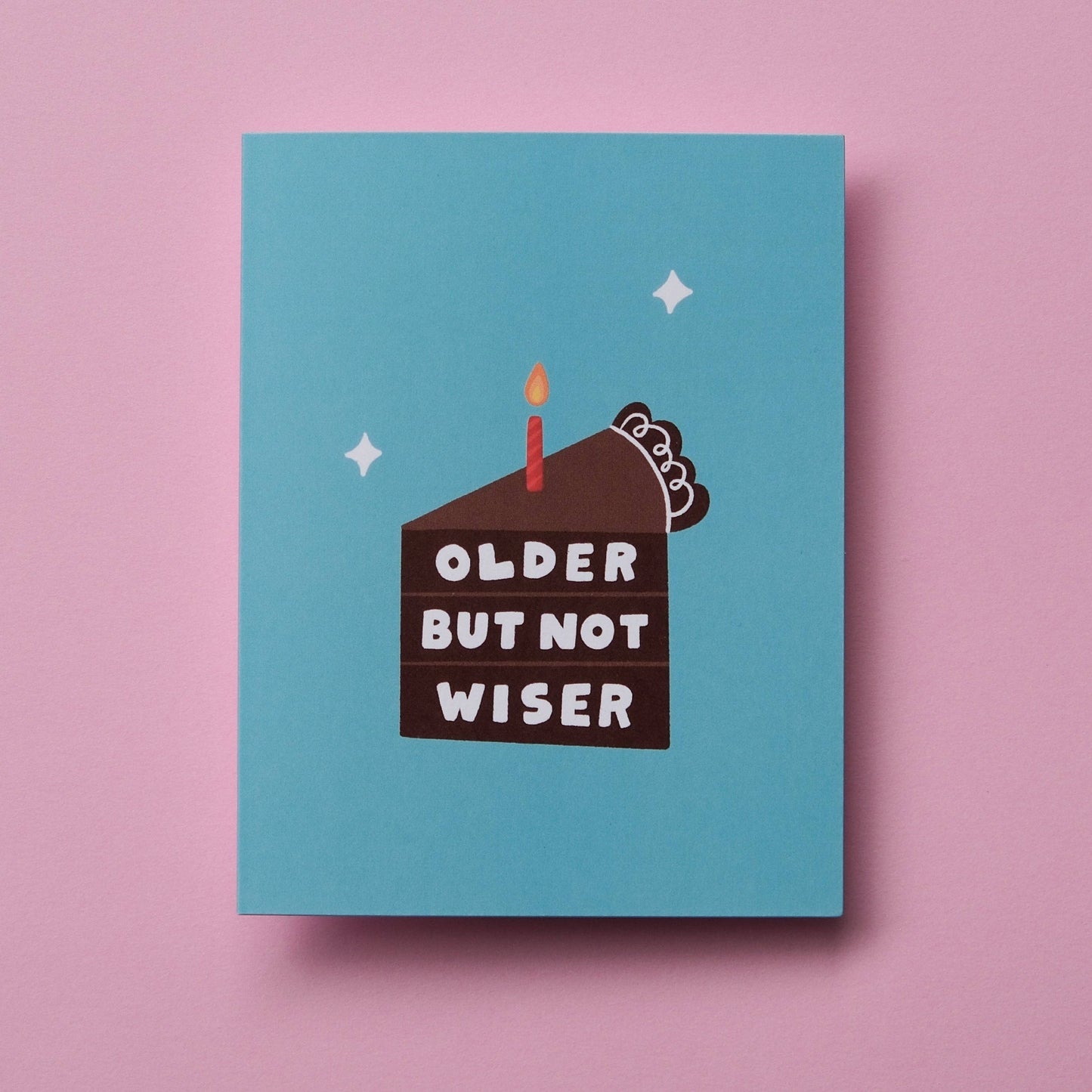 A square greeting card with a light blue background featuring a cartoon birthday cake with the text 'OLDER BUT NOT WISER' and a single candle, displayed on a solid pink background.