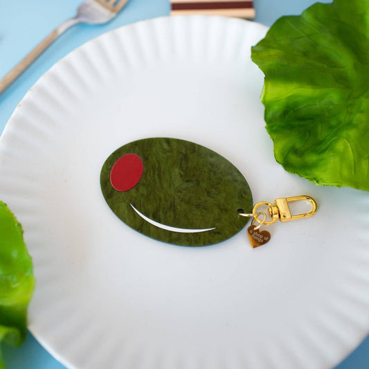Keychain shaped like a large green olive with red filling. Gold hardware/clasp. Shown on a white plate with fork on a blue tablecloth.