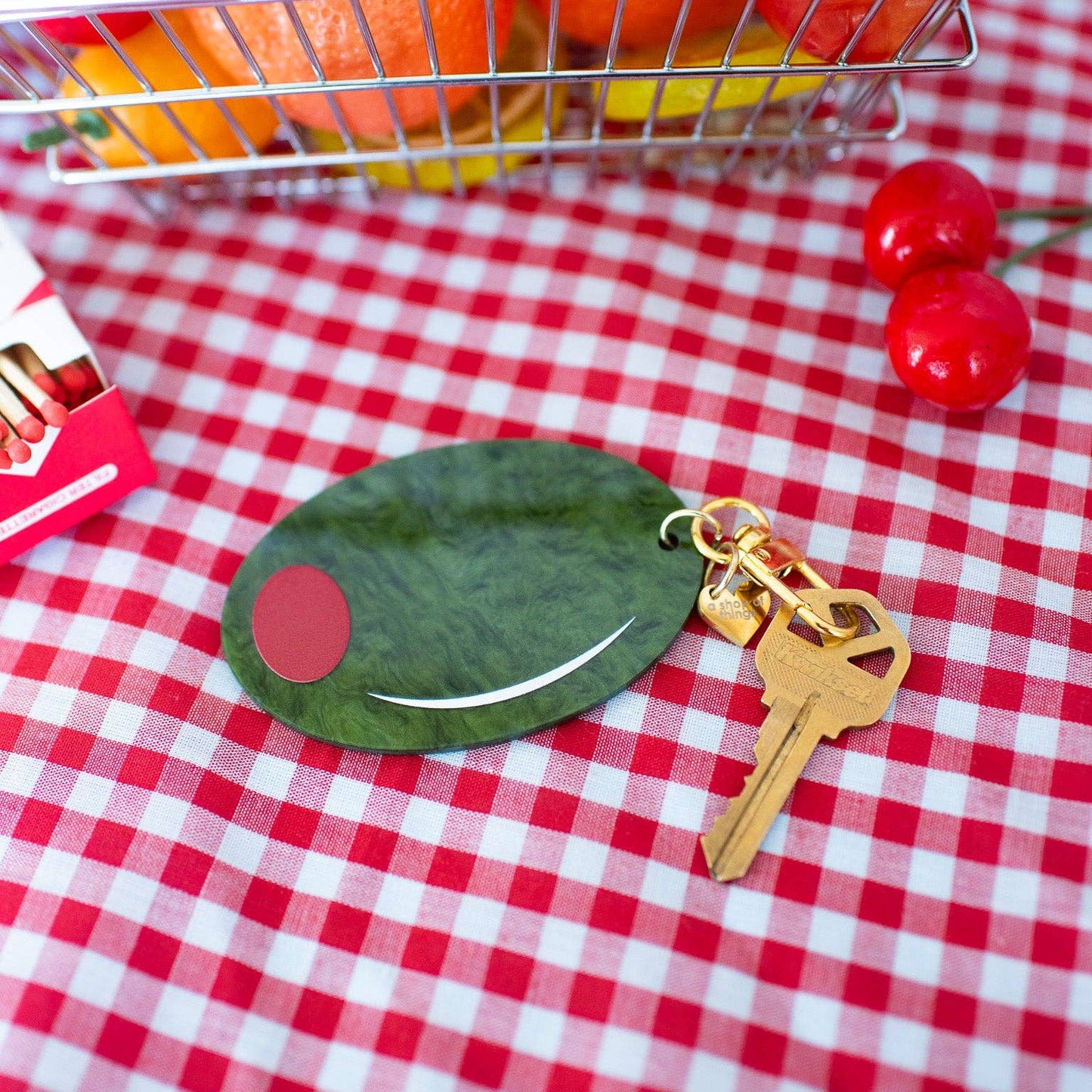 Keychain shaped like a large green olive with red filling. Gold hardware/clasp. Shown on a red and white checkered tablecloth.