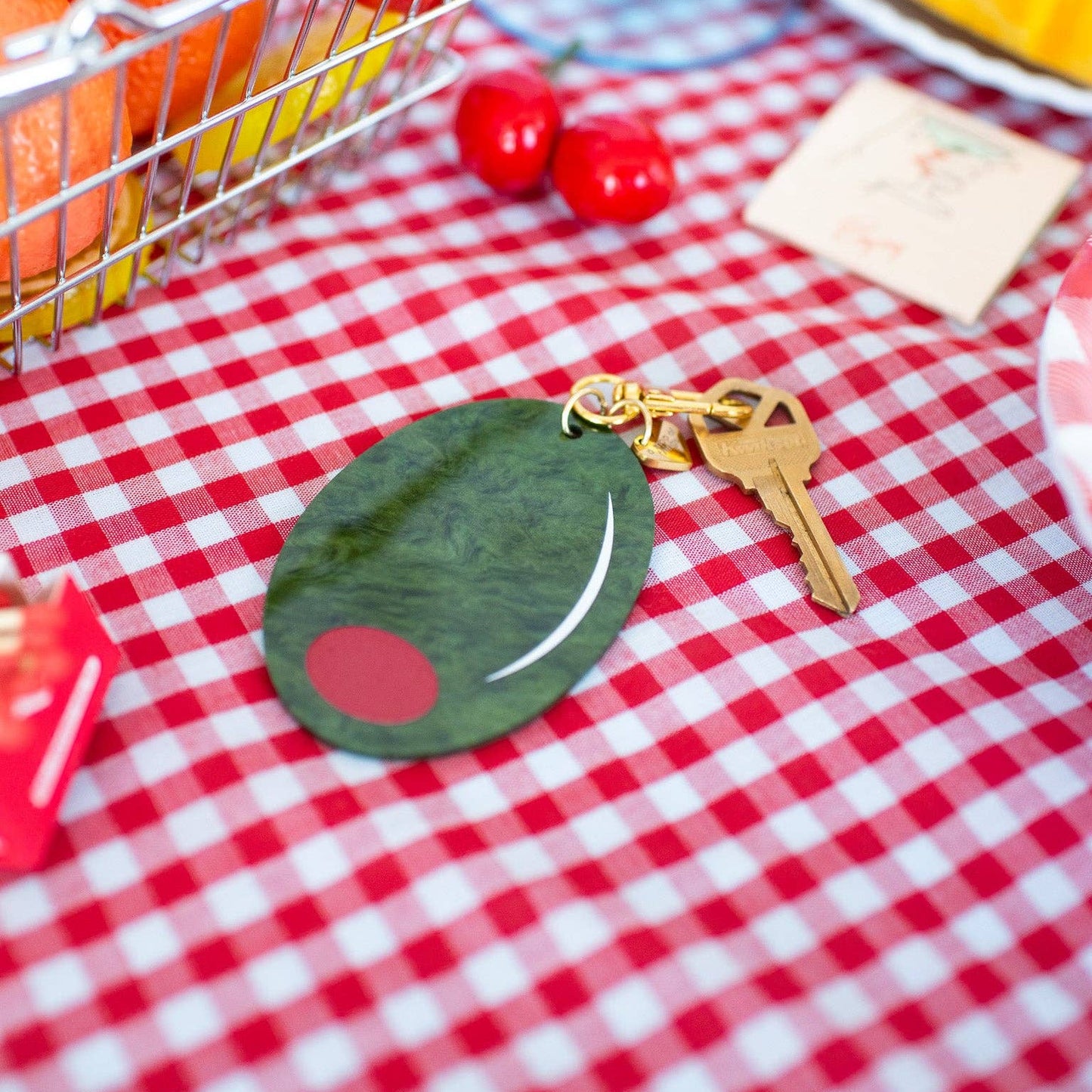Keychain shaped like a large green olive with red filling. Gold hardware/clasp. Shown on a red and white checkered tablecloth.
