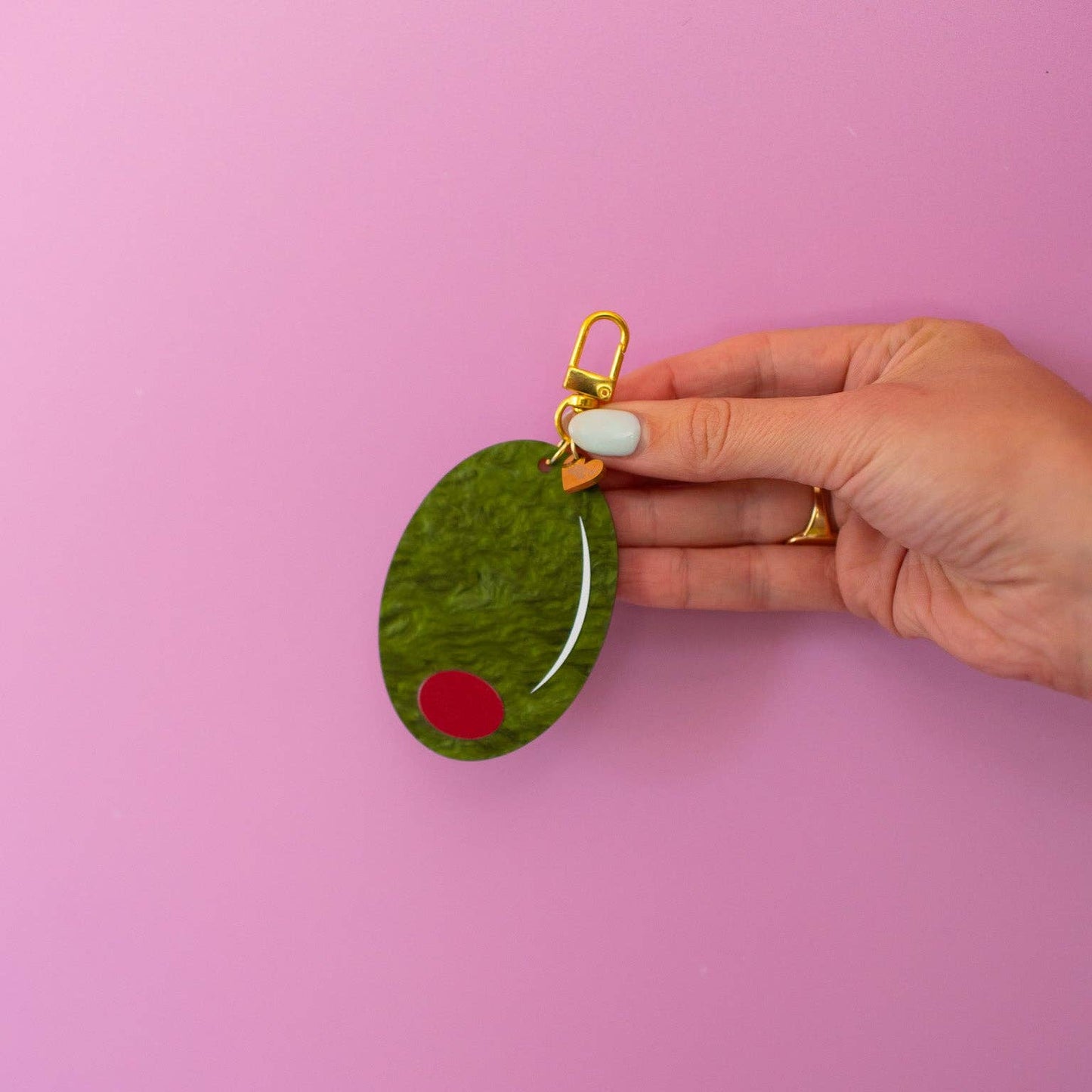 Keychain shaped like a large green olive with red filling. Gold hardware/clasp. Shown held in front of a pink background.