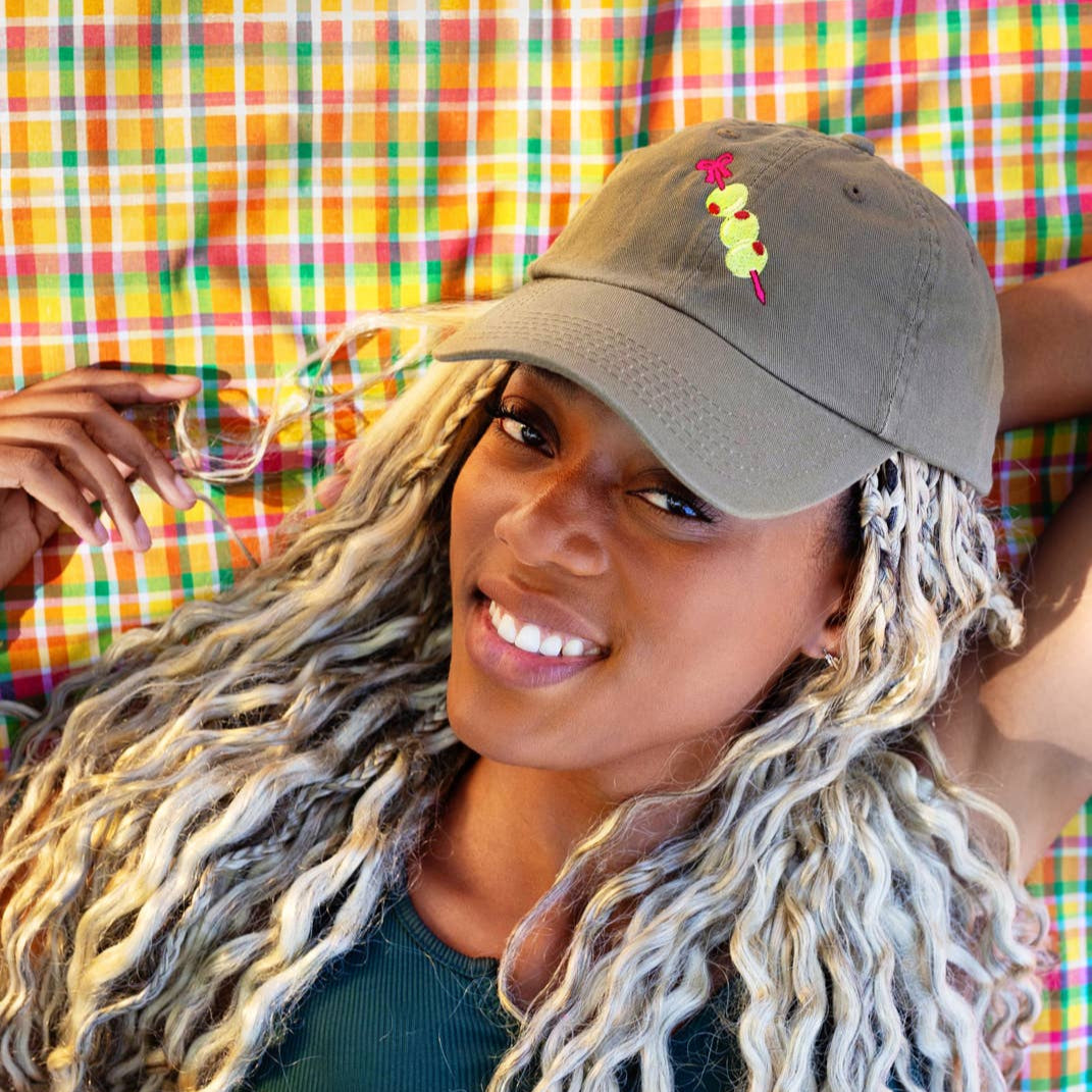 Grey baseball hat with embroidery detail of three stuffed green olives on a red cocktail pick. Shown on a smiling woman.