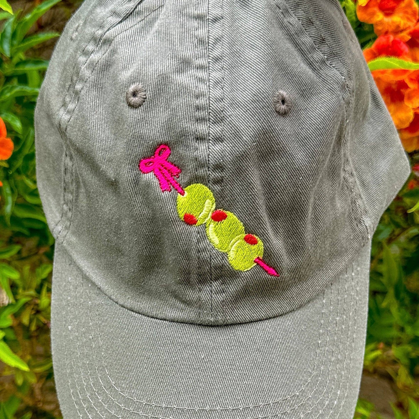 Close up photo: Grey baseball hat with embroidery detail of three stuffed green olives on a red cocktail pick.