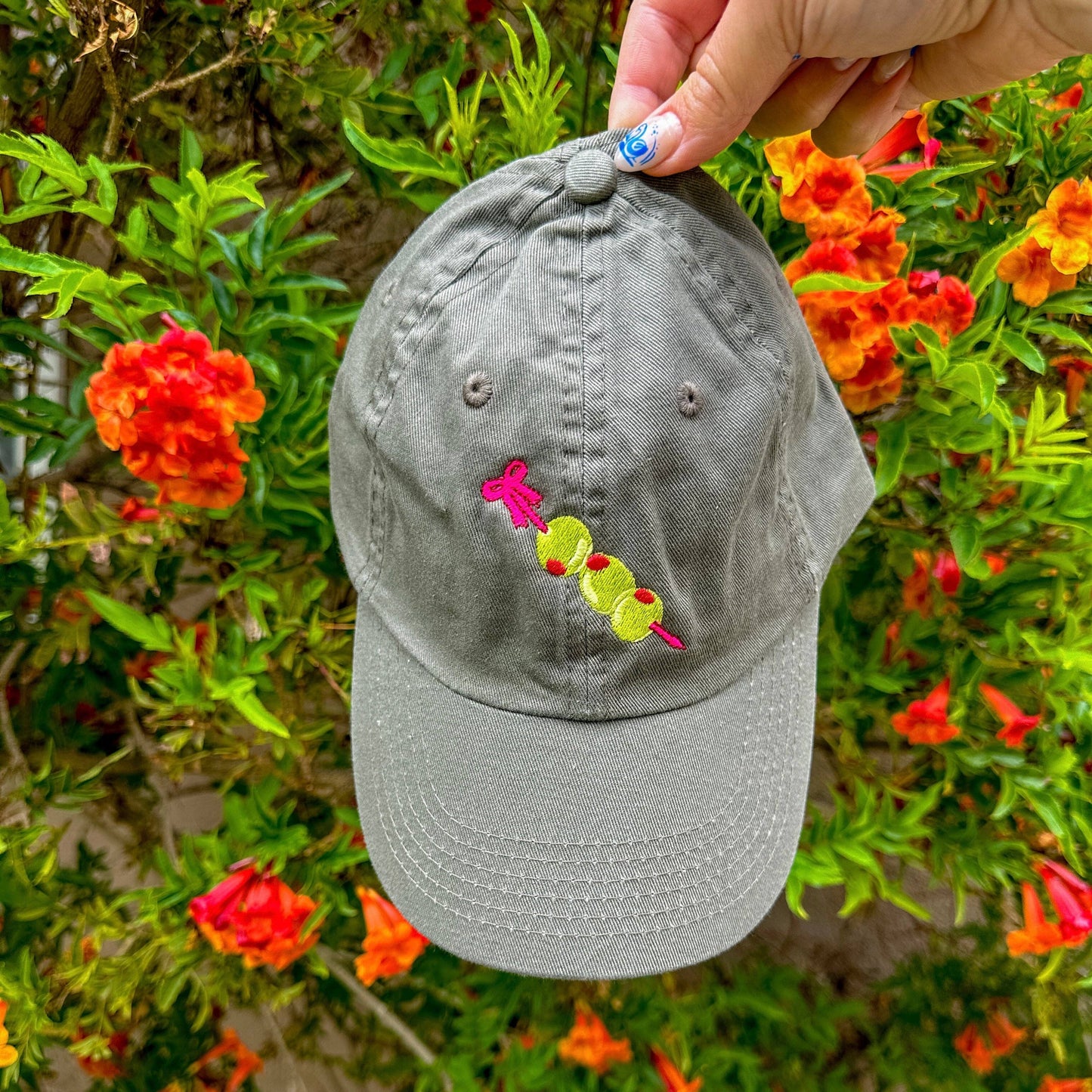 Close up photo: Grey baseball hat with embroidery detail of three stuffed green olives on a red cocktail pick.