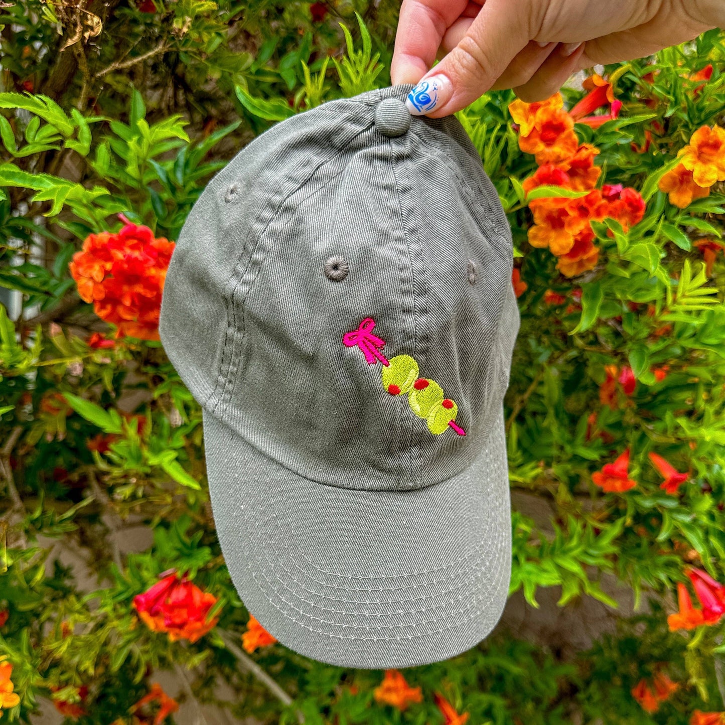 Close up photo: Grey baseball hat with embroidery detail of three stuffed green olives on a red cocktail pick.