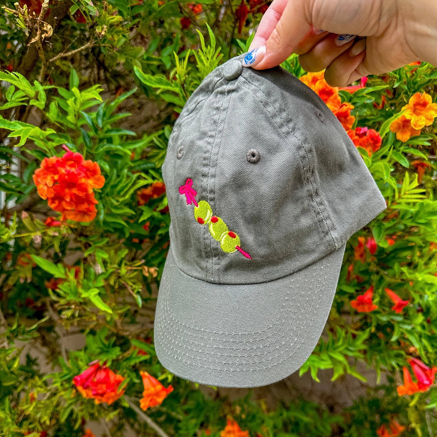 Close up photo: Grey baseball hat with embroidery detail of three stuffed green olives on a red cocktail pick.