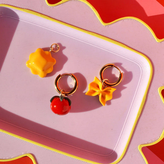 Pair of pasta earrings with yellow flower and red cherry design on a pink tray.