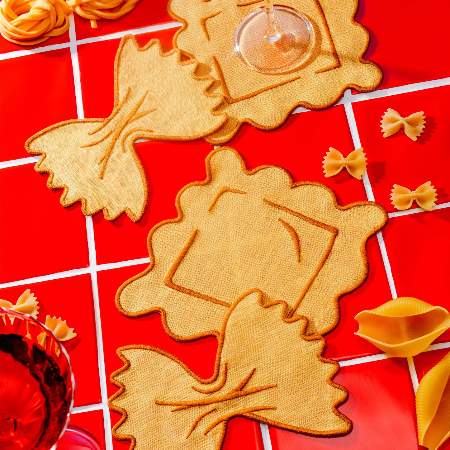 Linen pasta-shaped coasters on a red tiled surface with glasses of red wine.