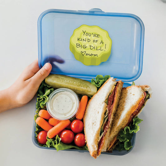 An open blue lunchbox held by a hand, filled with a sandwich, pickle, carrots, tomatoes, and a small cup of dressing. The lid has a yellow note that reads, 'You're kind of a big dill! mom'.