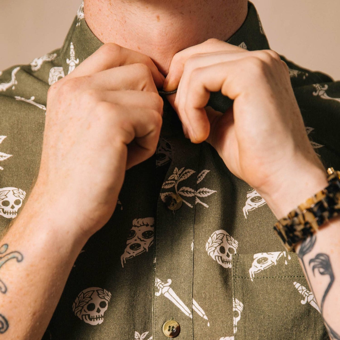 Man wearing a button up shirt that is oregano green with skulls, pizza and swords on it. He is adjusting the top button the collar.