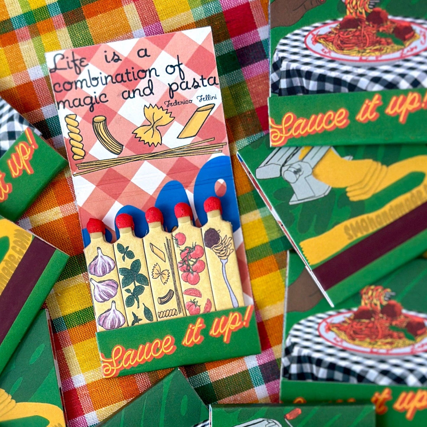 Spaghetti themed matchbook. Inside looks like red & white checkered tablecloth with pictures of different pasta shapes. Each match has a different image - garlic, basil, pasta shapes, tomatoes, spaghetti and meatball on a fork. Text on inner flap "Life is a combination of magic and pasta." Text on flap holding matches "Sauce it up!"