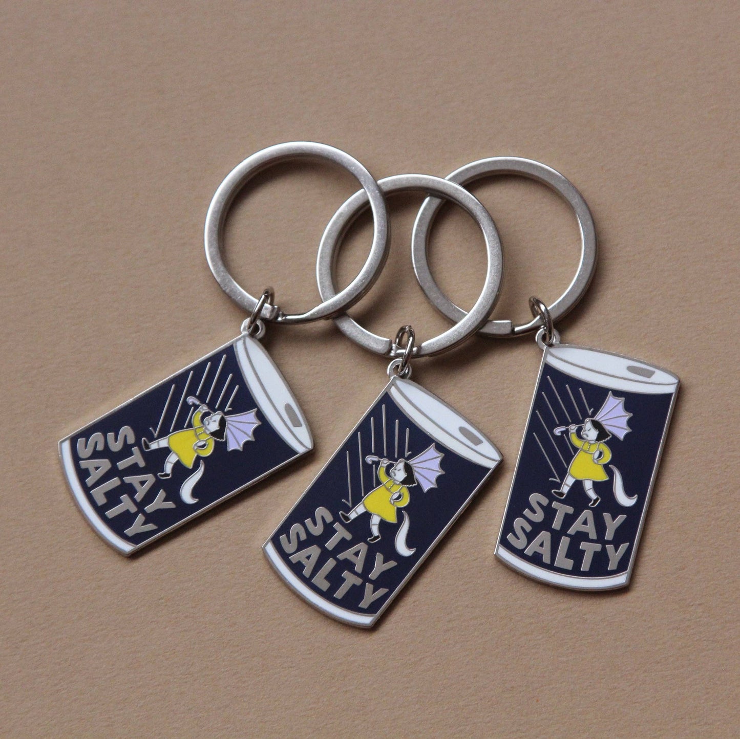 Three enamel keychains with silver metal rings and cylindrical charms featuring a navy blue background, a white cat in a yellow dress, and the text 'STAY SALTY' on a plain tan surface.