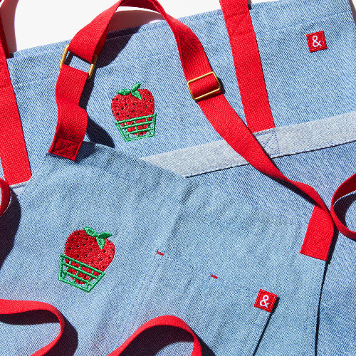 Blue apron with red straps. This apron is dedicated to strawberries with embroidery of strawberries in a basket and on a vine with little white flowers.
