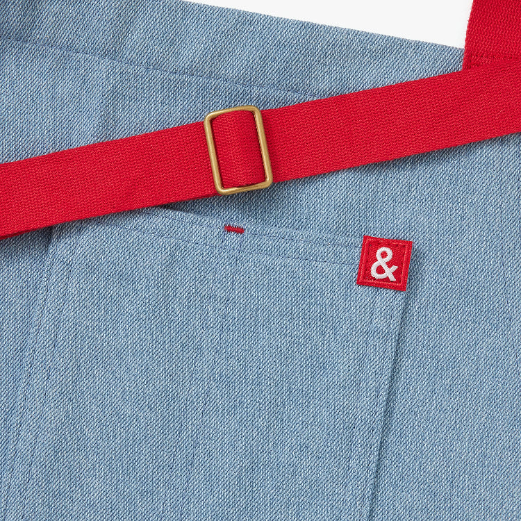 Blue apron with red straps. This apron is dedicated to strawberries with embroidery of strawberries in a basket and on a vine with little white flowers. Close up detail of red ampersand logo.
