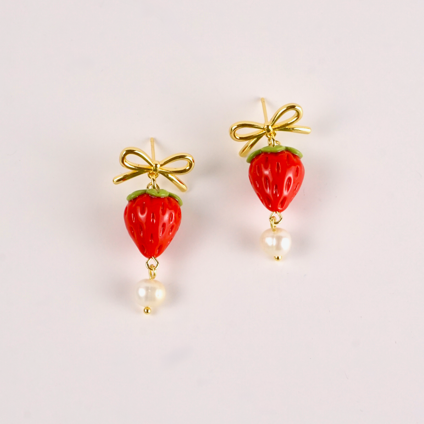 Strawberry-shaped earrings with gold bows and pearl drops on a light gray background