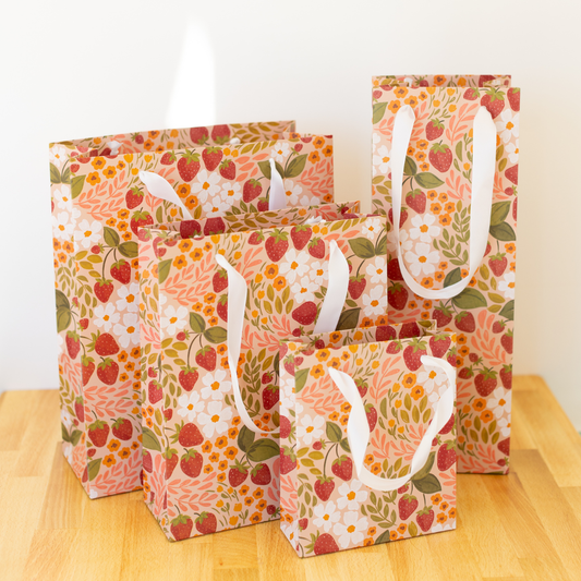 Gift bag shown in assorted sizes. Print on bag has a swirl of strawberries, leaves, and white/orange flowers on pale peach background. White fabric handles.
