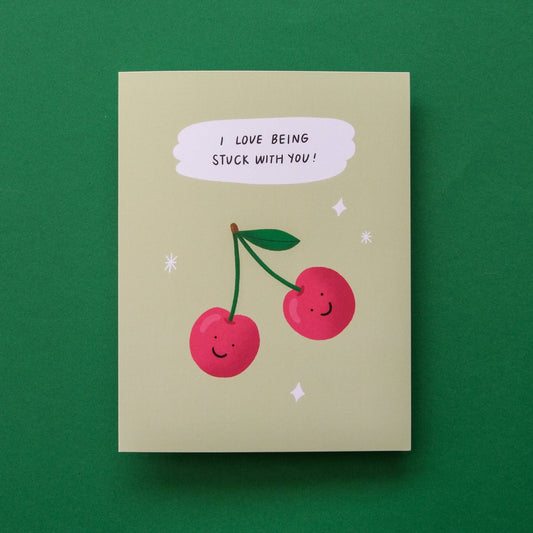 A square greeting card with a light beige background, featuring an illustration of two smiling red cherries with green stems and a speech bubble that reads 'I LOVE BEING STUCK WITH YOU!'. The card is photographed flat against a solid dark green background.