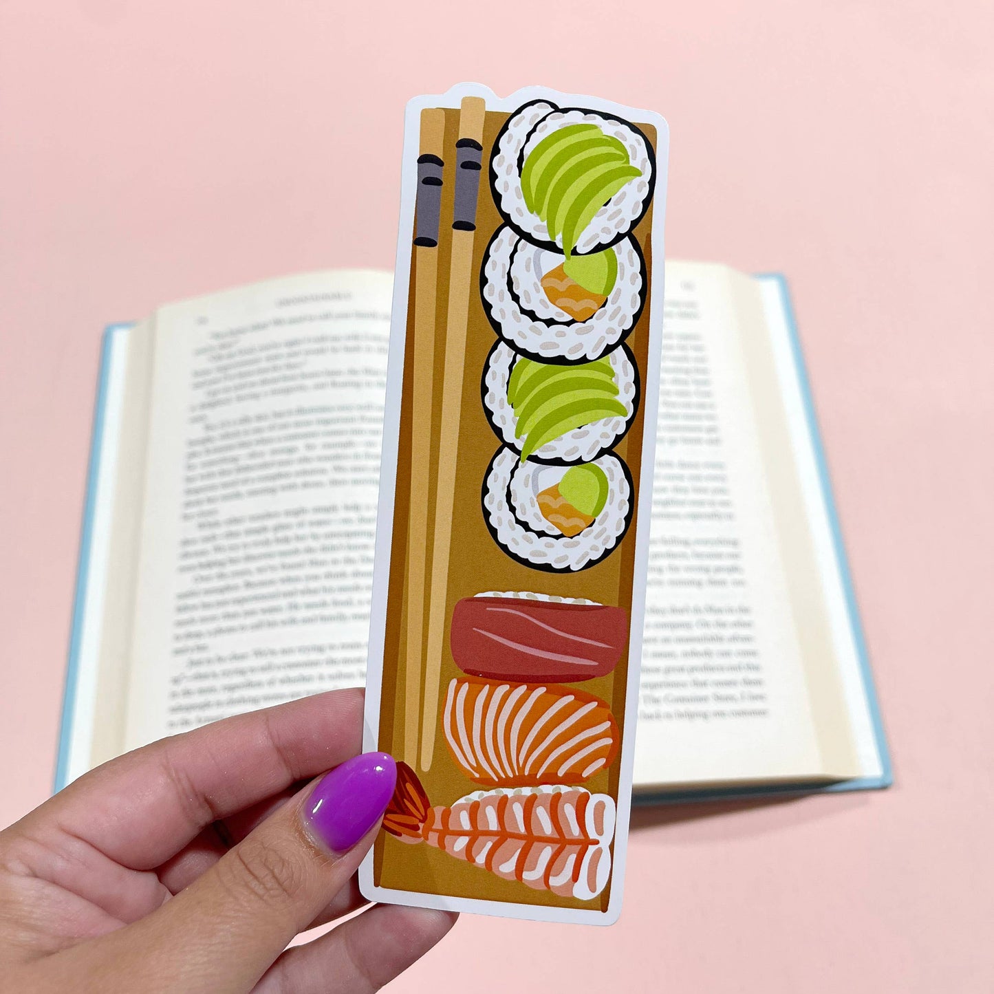 Hand holding a sushi-themed bookmark over an open book on a pink background
