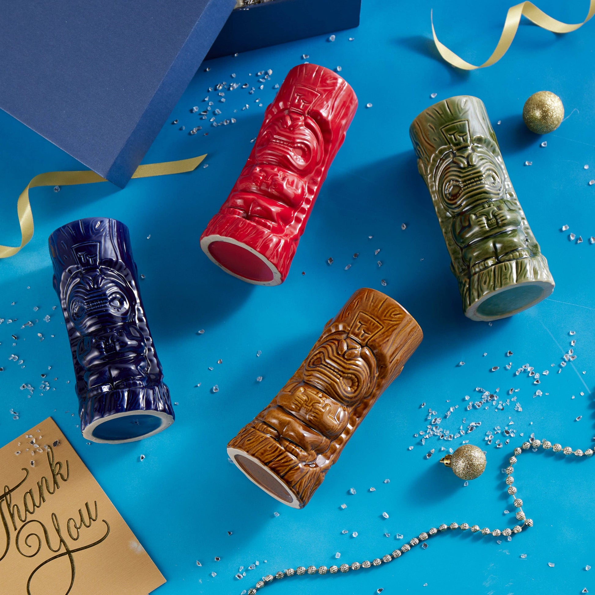 Four tiki mugs in blue, red, brown, and green on a blue background with decorative elements.