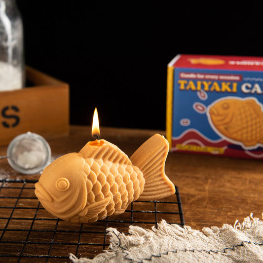 Fish-shaped candle with a lit wick on a cooling rack, with a box of Taiyaki in the background.
