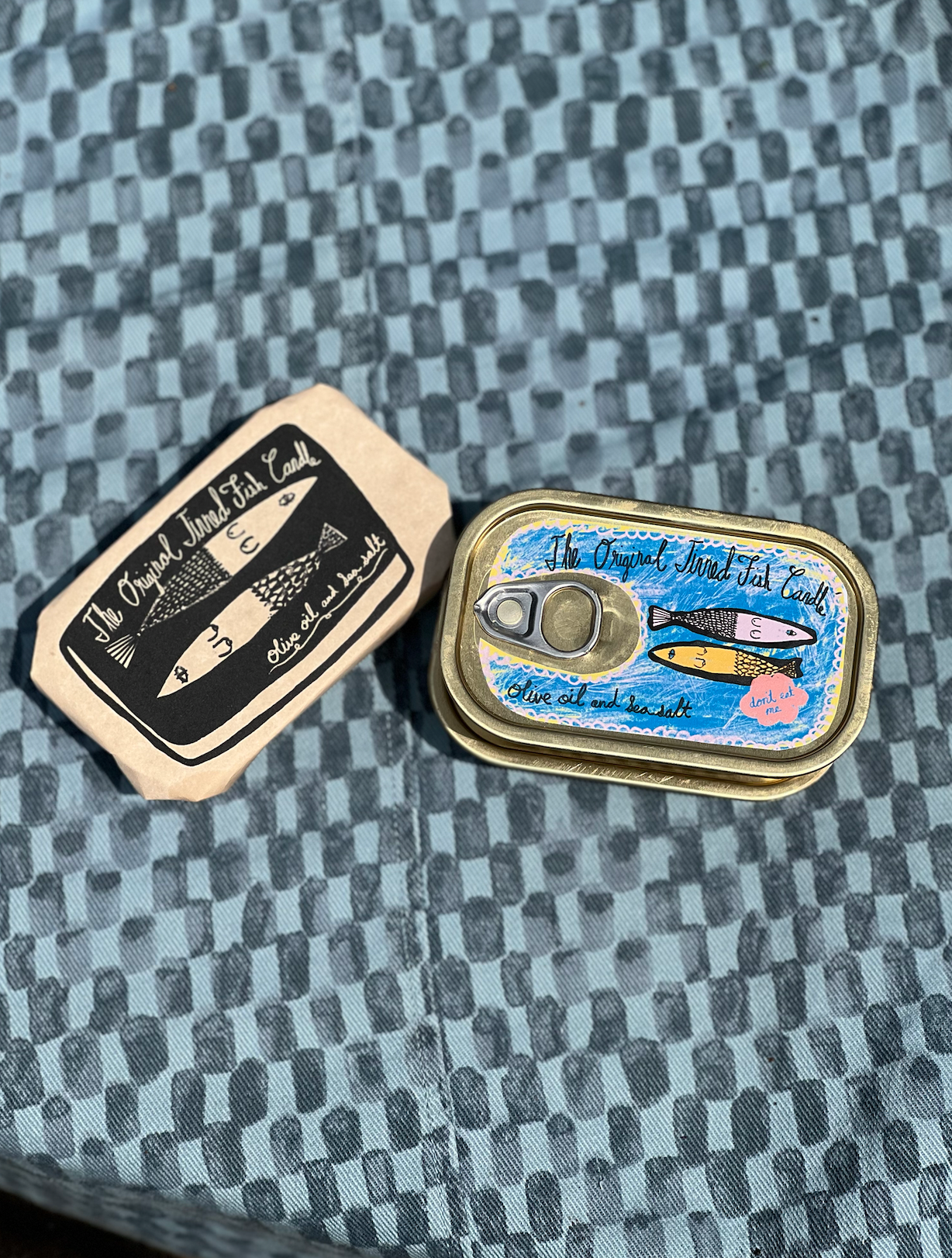 Outer paper packaging of the tinned fish candle next to the tinned fish can it comes in