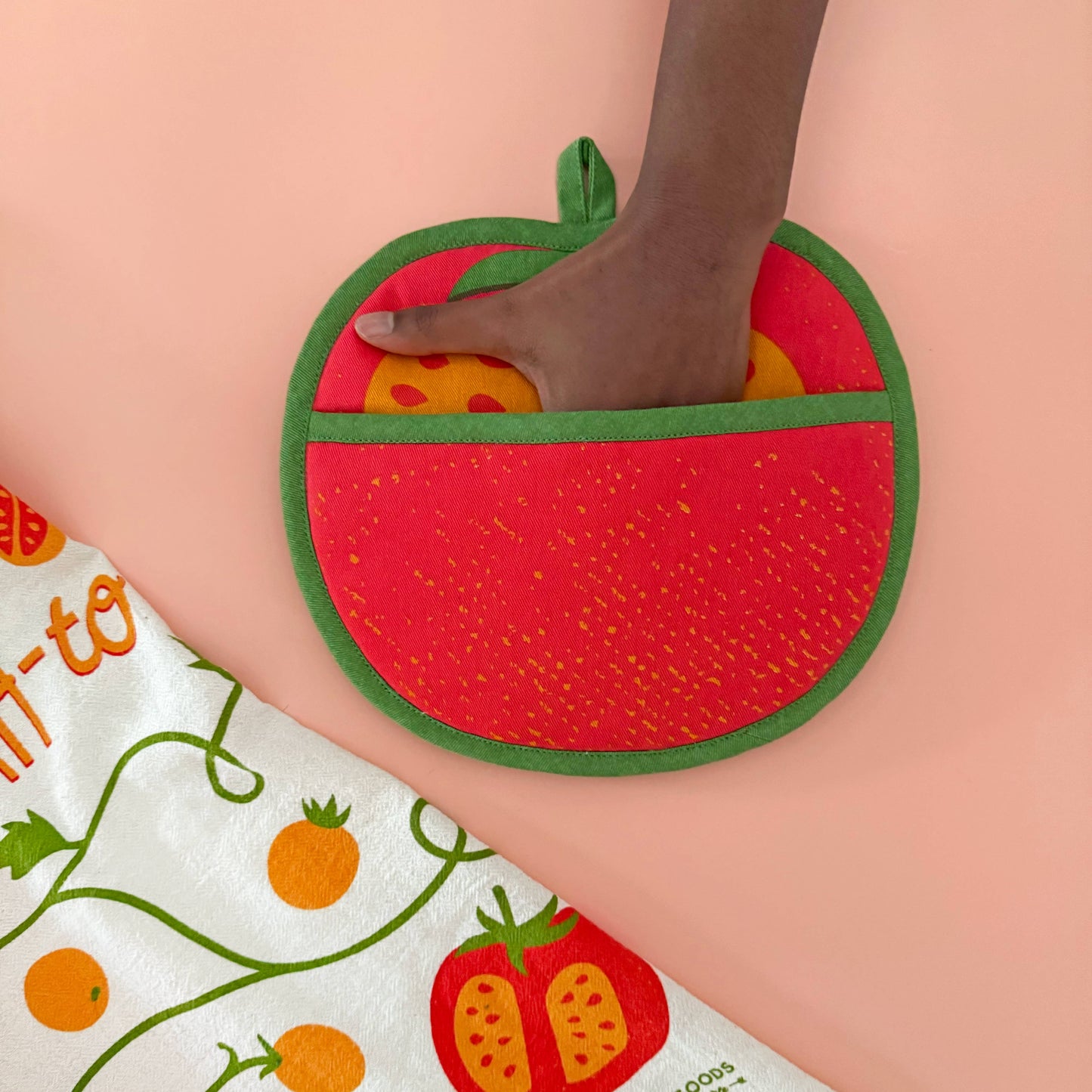 Hand holding a red tomato-shaped pot holder with a green border on a light pink background.
