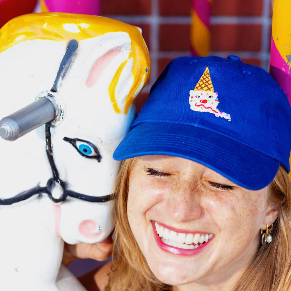 An upside down ice cream cone embroidered onto a blue baseball hat. Ice cream cone looks like: scoop of strawberry ice cream on a sugar cone with rainbow sprinkles, two chocolate chips for eyes, a maraschino cherry for a nose, and a sweet licorice rope smile. Shown on a smiling woman.
