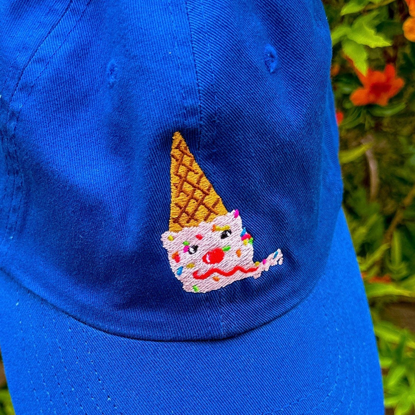 An upside down ice cream cone embroidered onto a blue baseball hat. Ice cream cone looks like: scoop of strawberry ice cream on a sugar cone with rainbow sprinkles, two chocolate chips for eyes, a maraschino cherry for a nose, and a sweet licorice rope smile. Detail photo of ice cream embroidery.