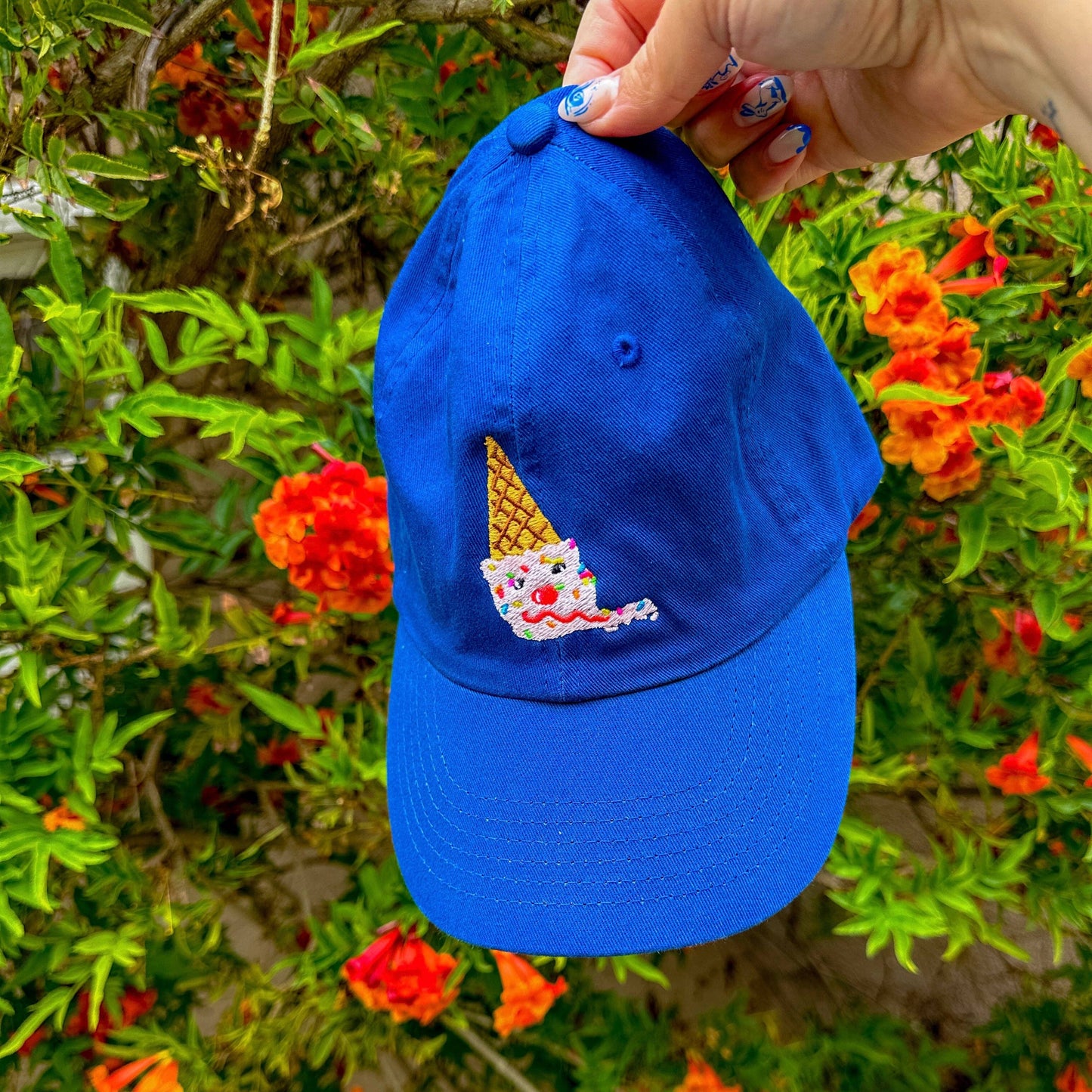 An upside down ice cream cone embroidered onto a blue baseball hat. Ice cream cone looks like: scoop of strawberry ice cream on a sugar cone with rainbow sprinkles, two chocolate chips for eyes, a maraschino cherry for a nose, and a sweet licorice rope smile. Shown held in front of a flowering bush.