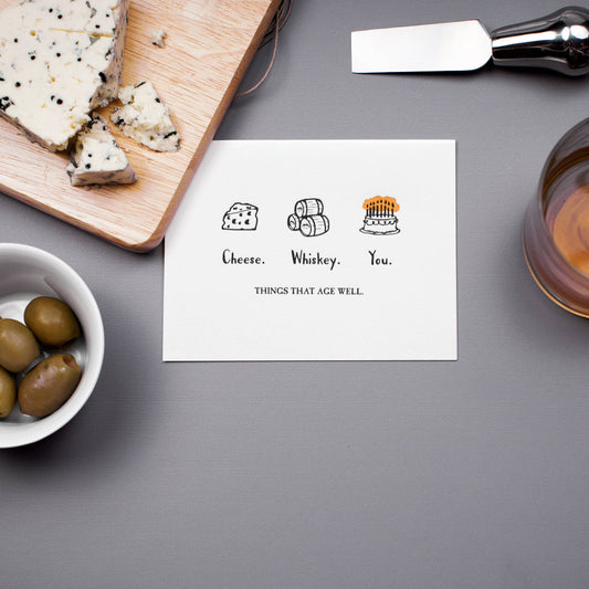 Greeting card with illustrations and text 'Cheese, Whiskey, You. Things that age well.' on a table with cheese and olives.
