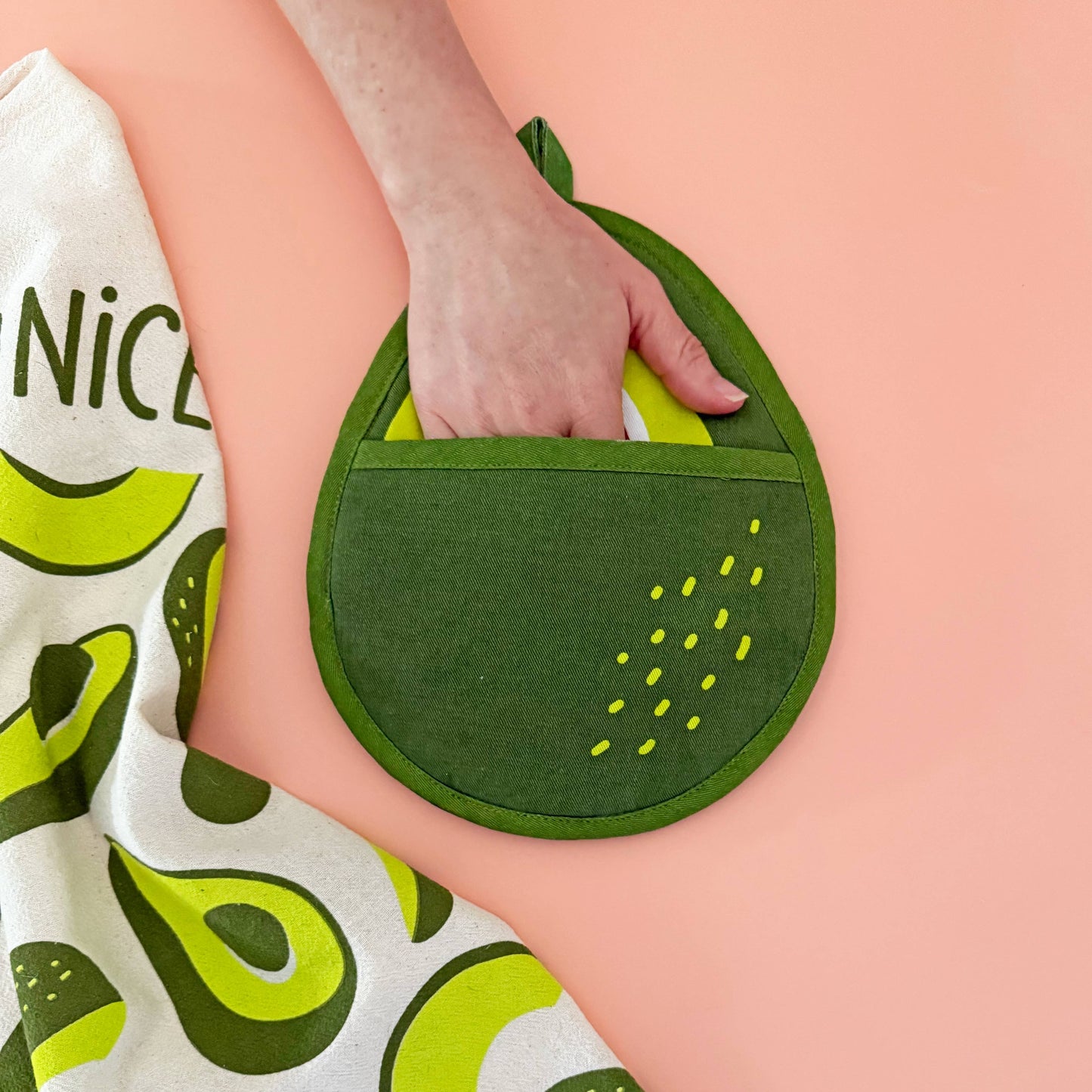 Green pot holder held by a hand on a pink background with a white towel featuring green avocado prints.