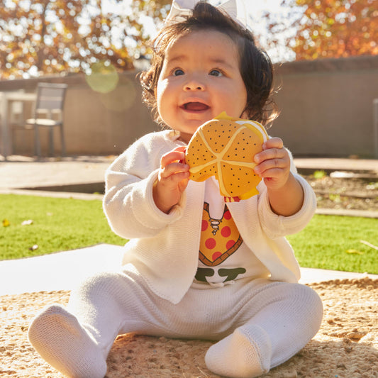 Child holding a yellow teether toy outdoors with trees in the background