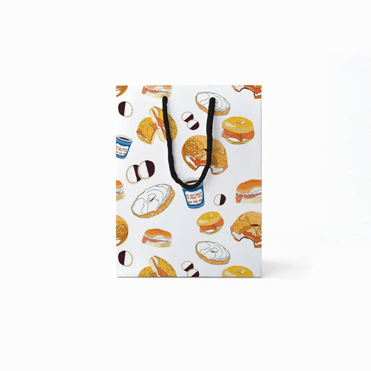 Gift bag with black rope handles and designed with various illustrations of black & white cookies, coffee in a blue paper cup, bagels with schmear or lox