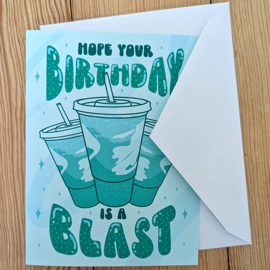 Birthday card with a Baja Blast soda graphic and text on a wooden surface with white envelope