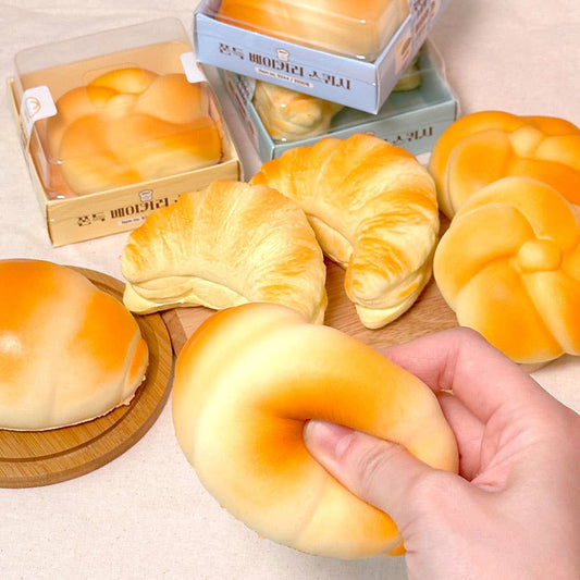 Hand holding a croissant-shaped stress ball with other bread items and packaging in the background.