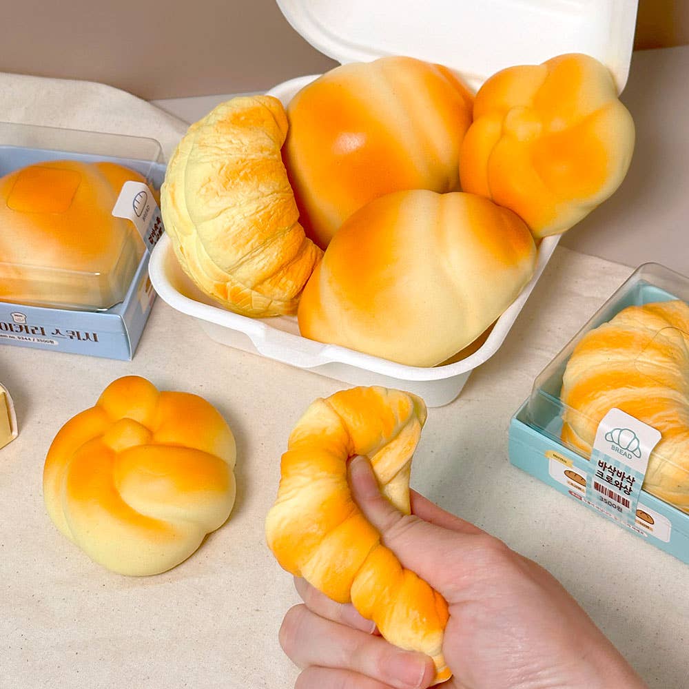 Stress balls that look like baked goods in a white container with a hand holding a croissant stress ball on a light surface.