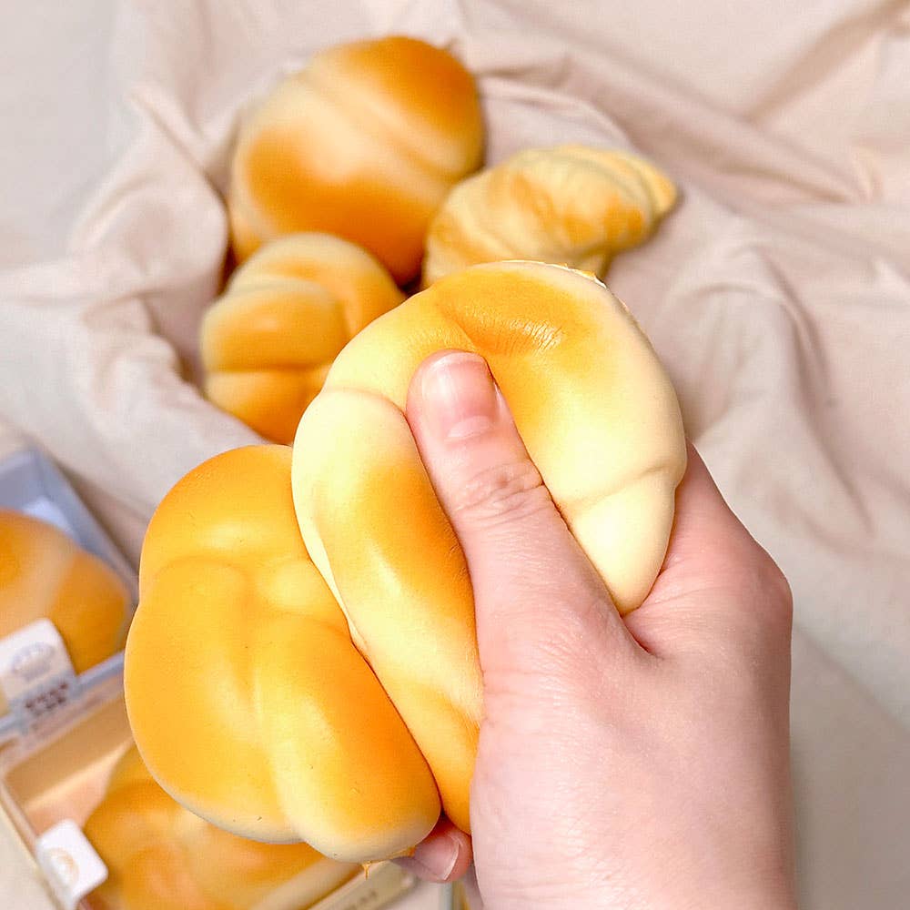 Hand holding a soft, yellow stress ball that looks like bread with more toys and a box in the background.