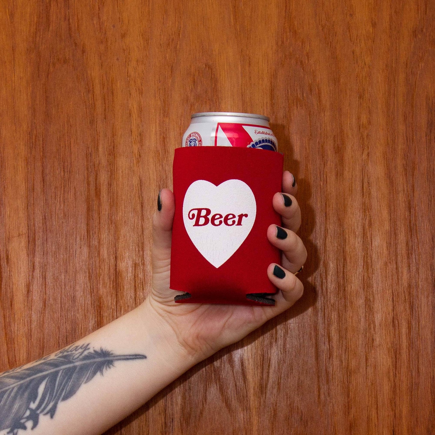 Hand holding a can of beer with a red sleeve labeled 'Beer' against a wooden background