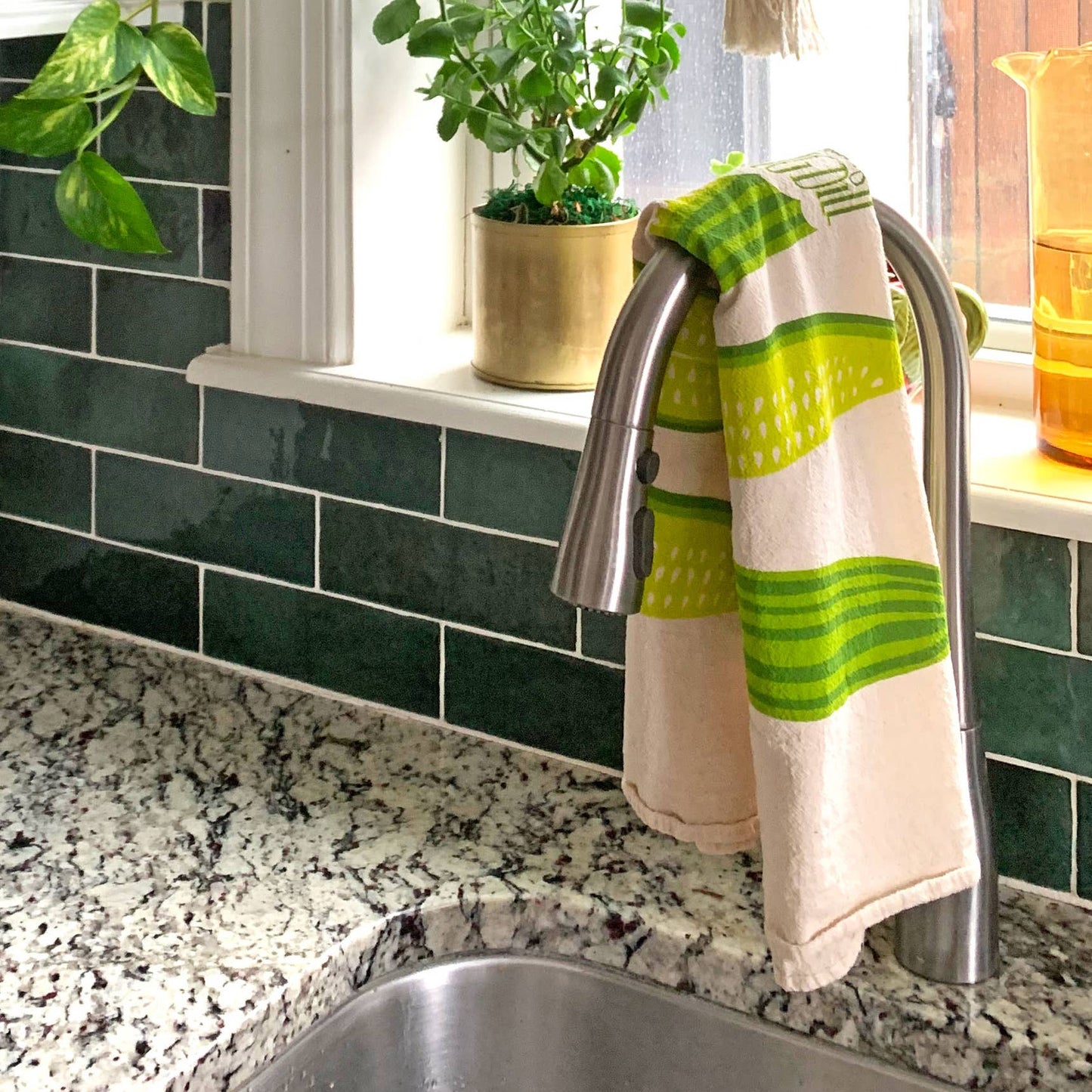 Kitchen sink area with the dill pickle spear towel hanging on the faucet, green tiles, and a plant.