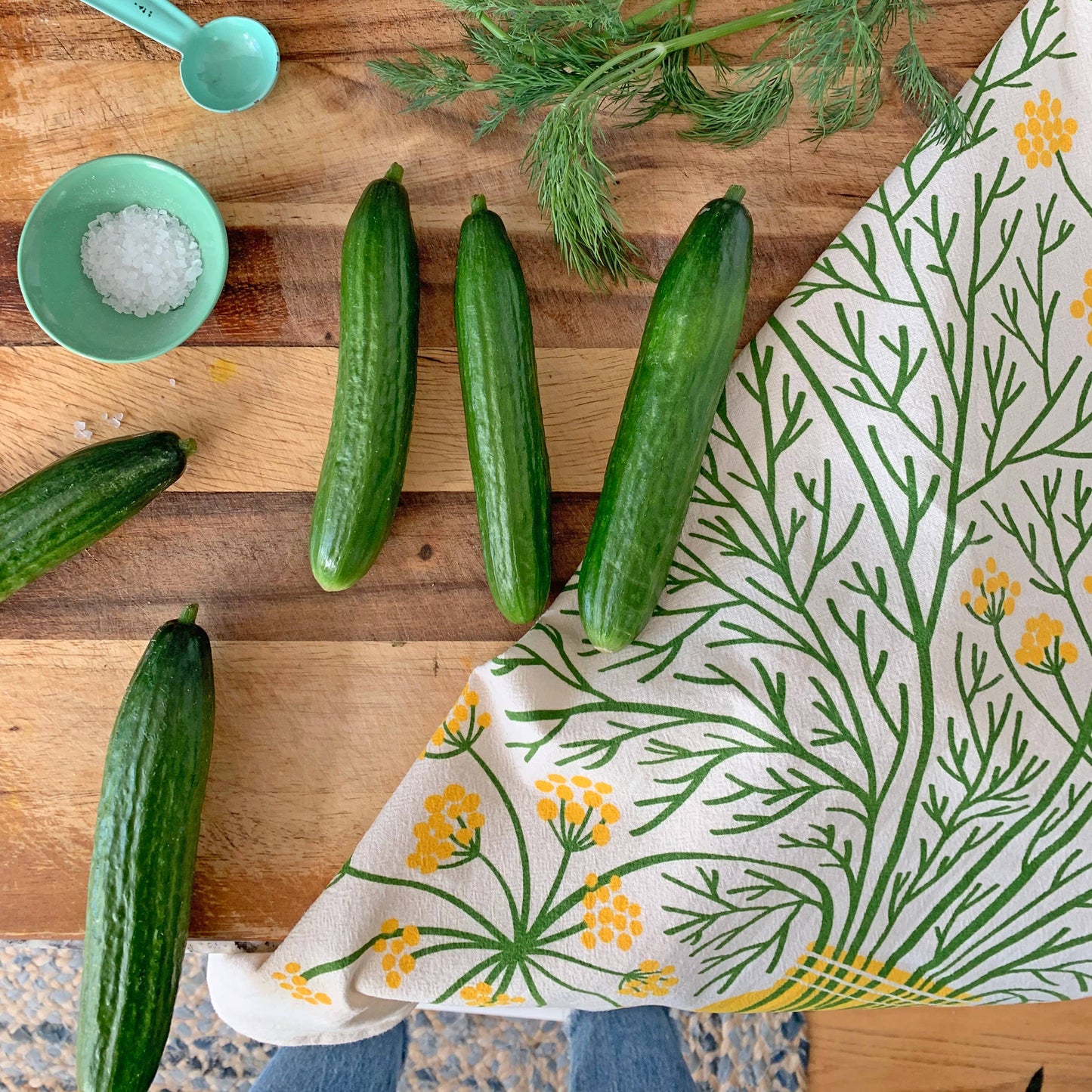 Cucumbers on a wooden cutting board with a dill floral towel and kitchen items.