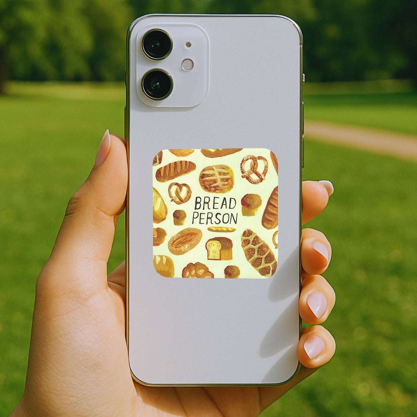 A hand holds a silver smartphone outdoors, showing a square sticker on the back with a pattern of various breads and the text 'BREAD PERSON'. The background is a blurred green park with trees.