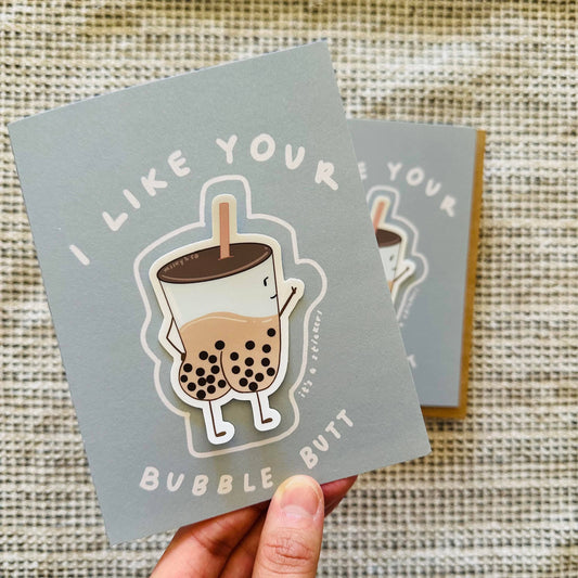 Greeting card that reads "I like your bubble butt" accompanied with a sticker of an illustrated boba cup with a butt filled with tapioca balls and milk tea looking back at it