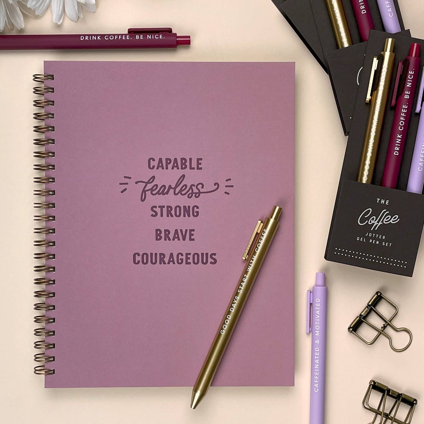 Purple notebook with motivational quote, pens, and flowers on a light background