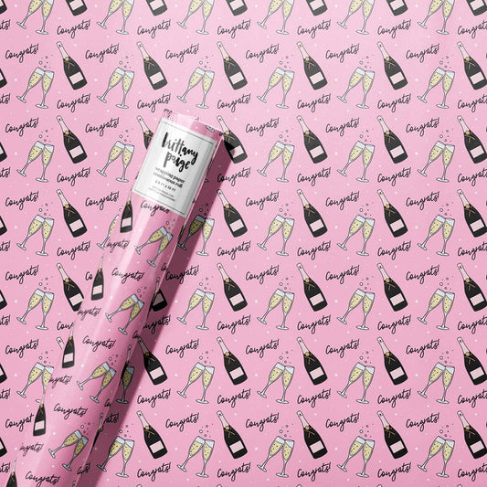 roll of wrapping paper that has a pink background with a continuous pattern of a champagne bottle, two flutes clinking and the text "congrats" all over