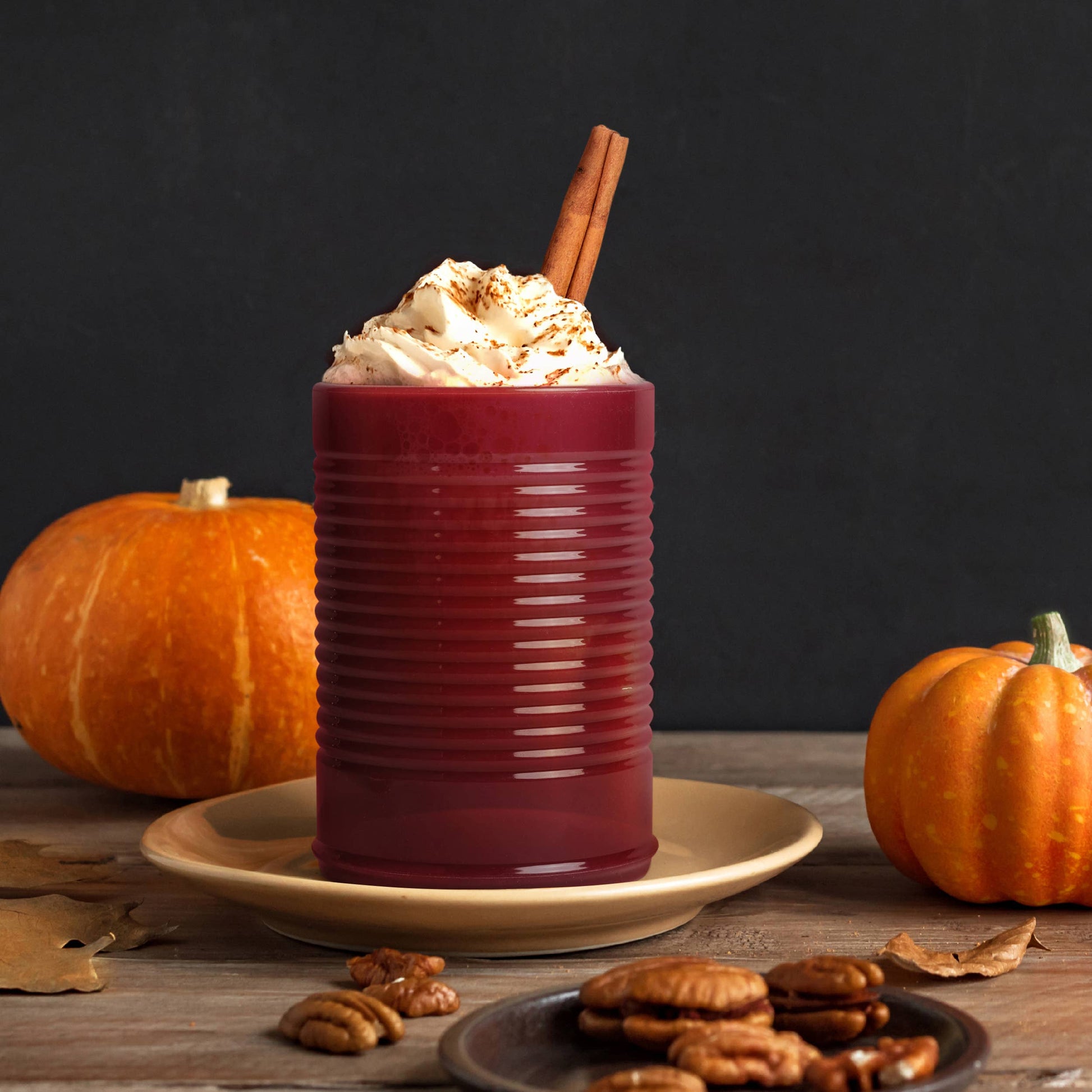 Pumpkin spice latte in a red, cranberry sauce silicone cup with whipped cream and a cinnamon stick, surrounded by pumpkins and pecans on a wooden surface.