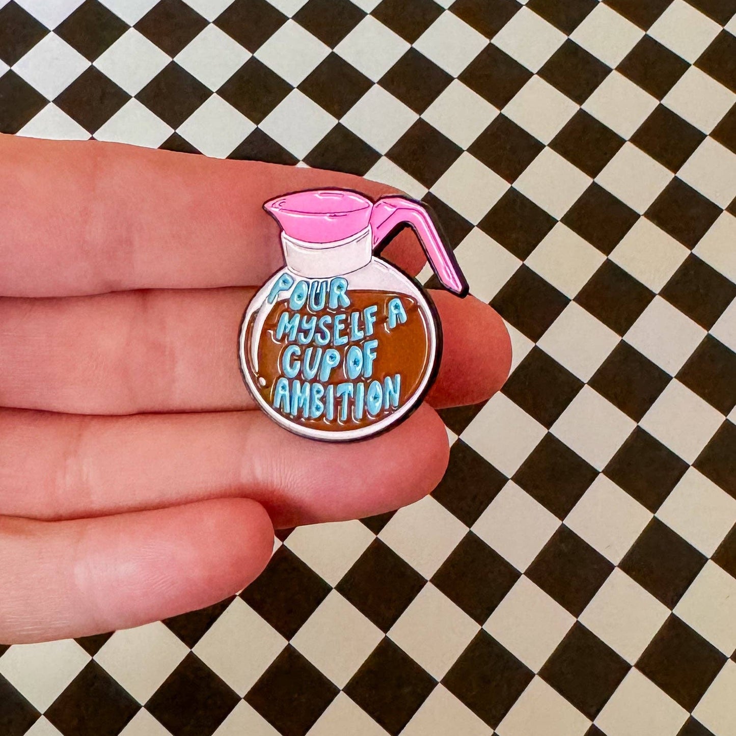 Enamel pin of a coffee pot with 'Pour myself a cup of ambition' text held by a hand on a checkered background