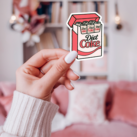 Hand holding a Diet Coke box-shaped sticker with a blurred indoor background